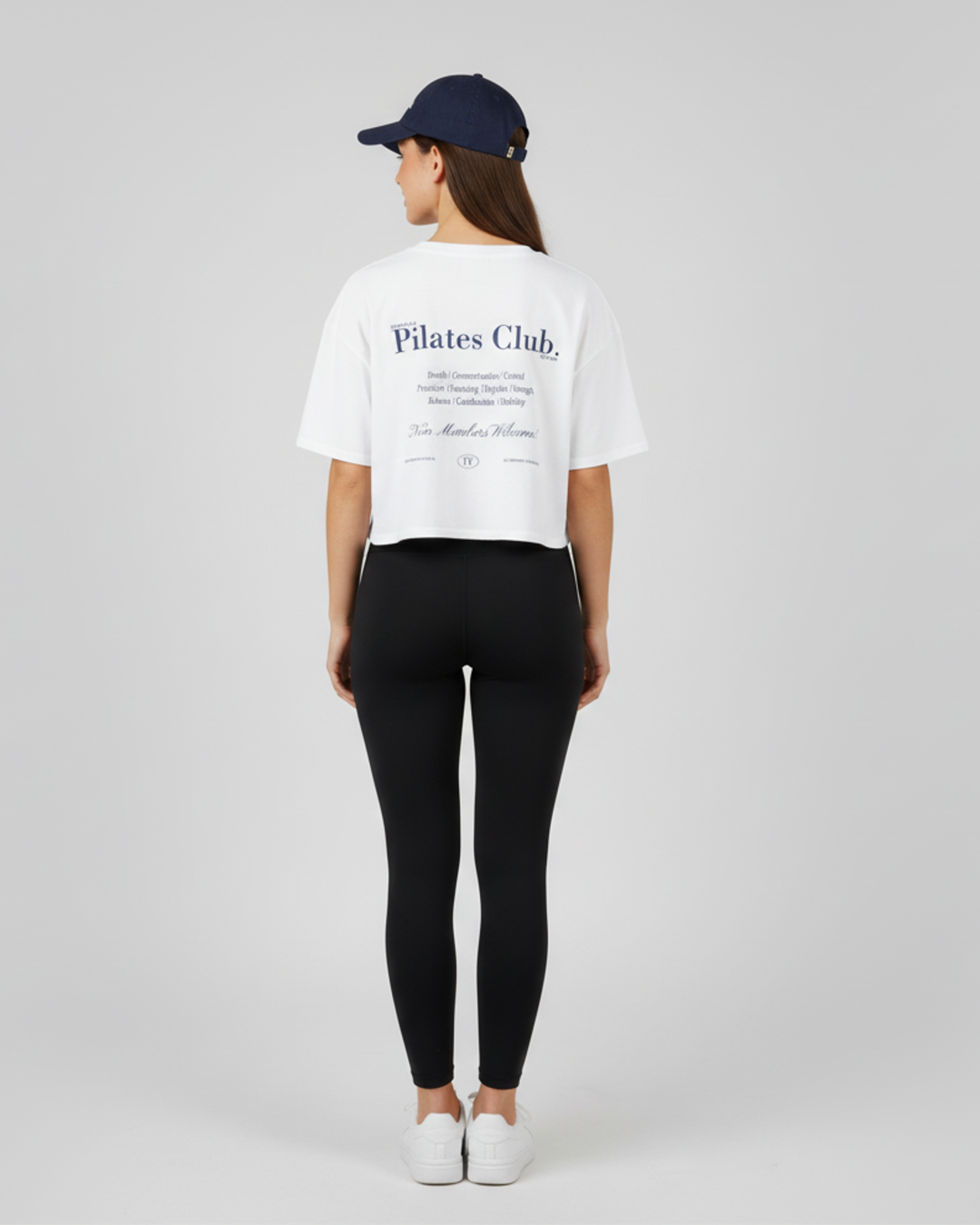 Person wearing a white 'Pilates Club' t-shirt, black leggings, and a navy cap on a light gray background