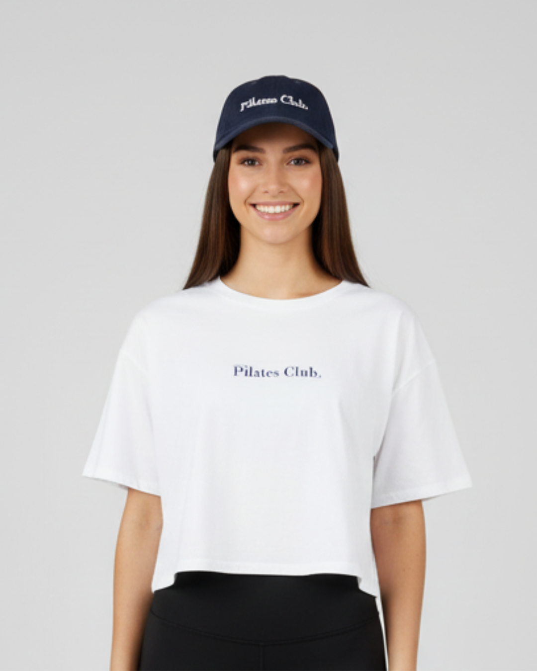 Woman wearing a white 'Pilates Club' t-shirt and navy cap on a light gray background