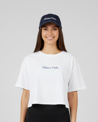 Woman wearing a white 'Pilates Club' t-shirt and navy cap on a light gray background