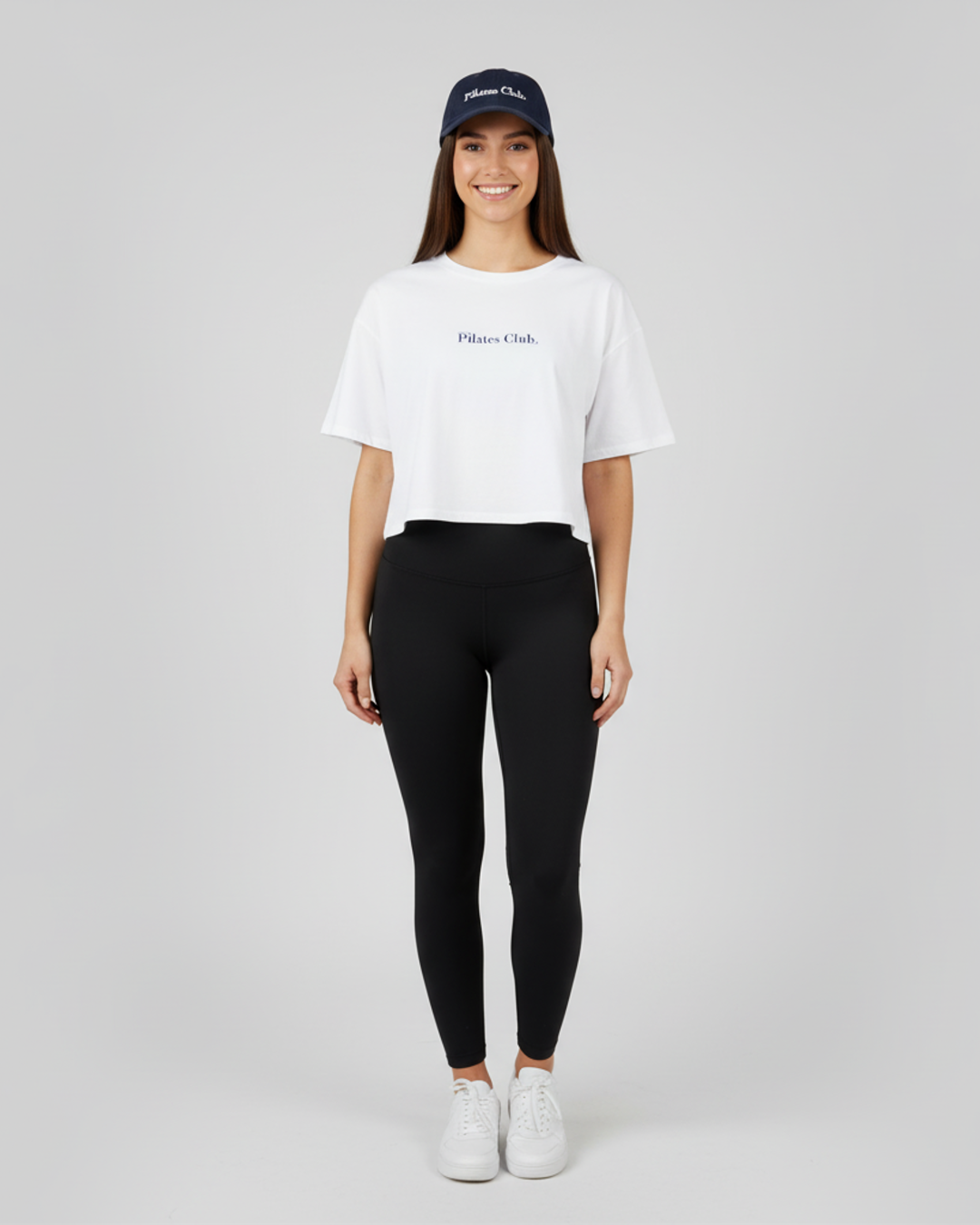 Woman wearing a white t-shirt, black leggings, and a navy cap on a light gray background